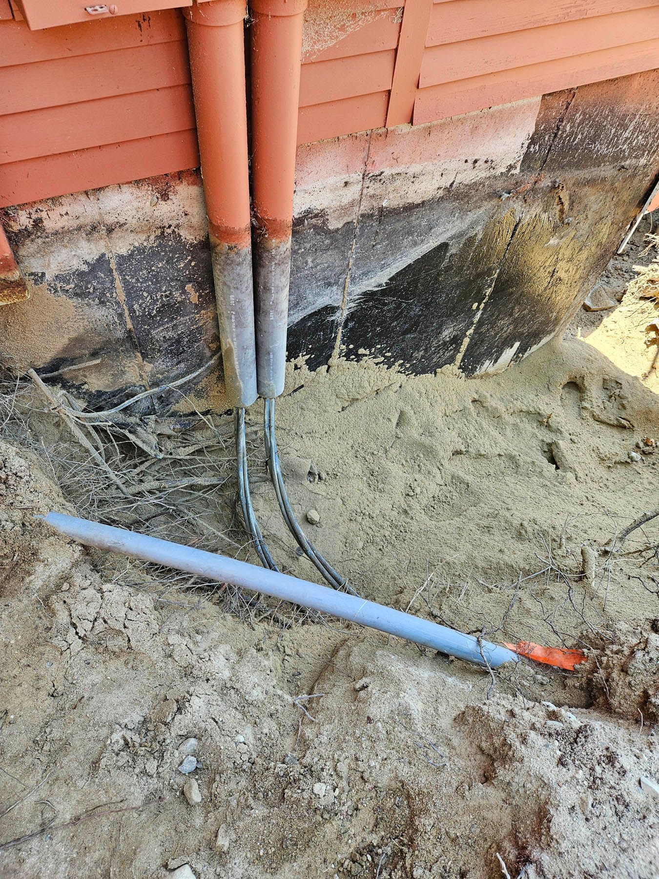 Pipes emerge from a building's foundation, buried in sand. One blue, two brown.