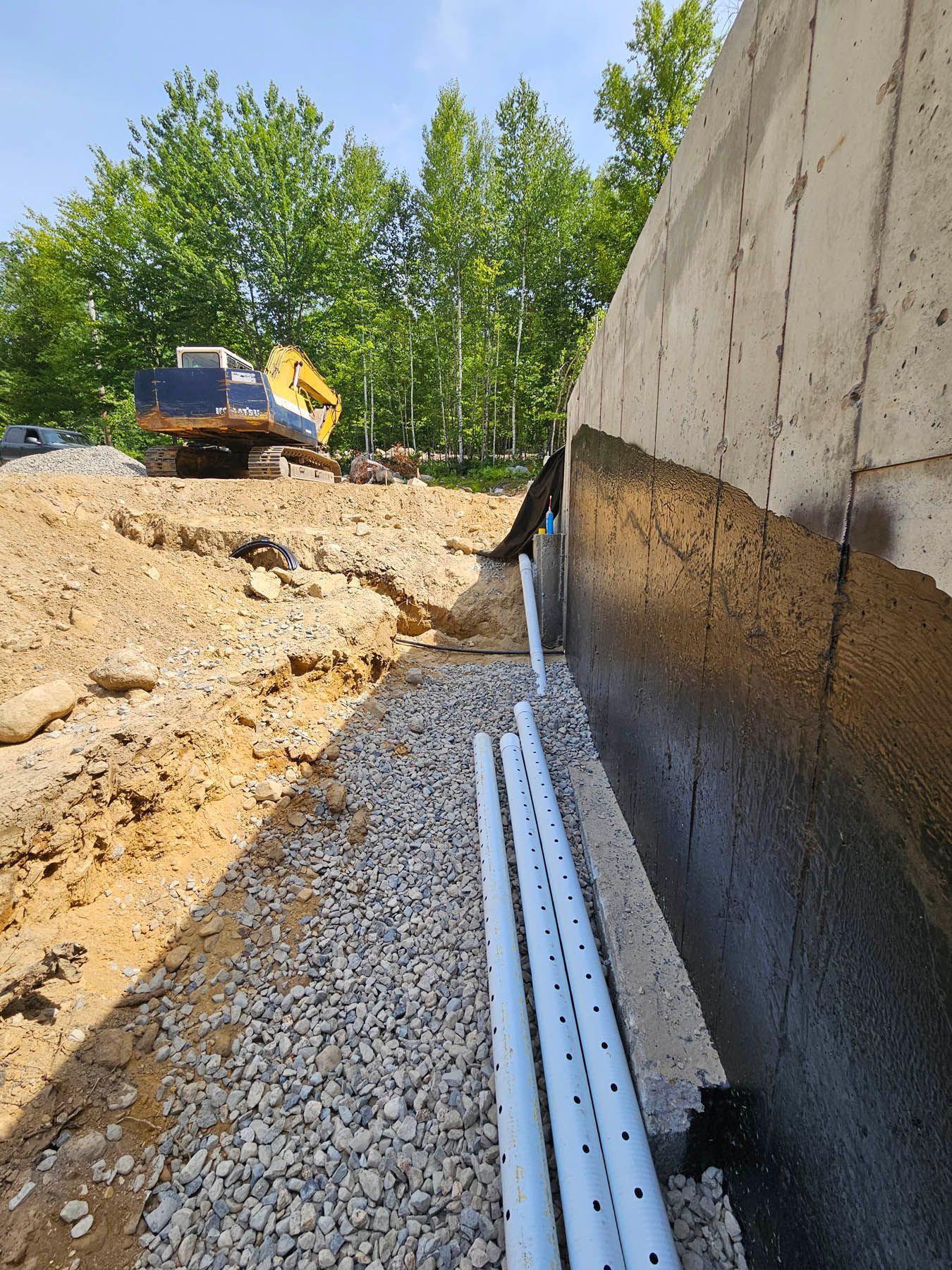 Construction site with a concrete wall, drain pipes, gravel, and an excavator, outdoors on a sunny day.