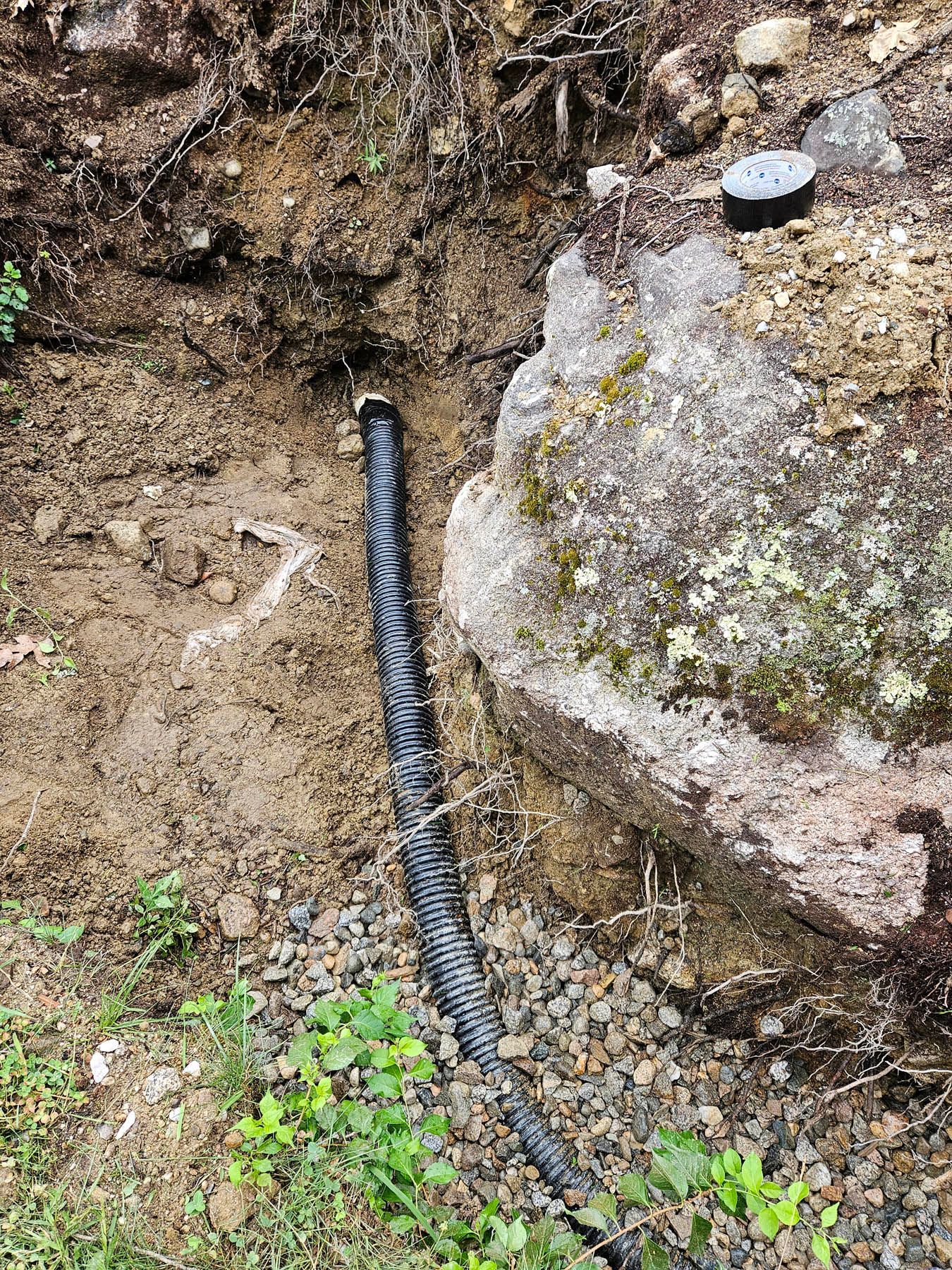 Black corrugated pipe in a trench next to a large rock in an outdoor setting.