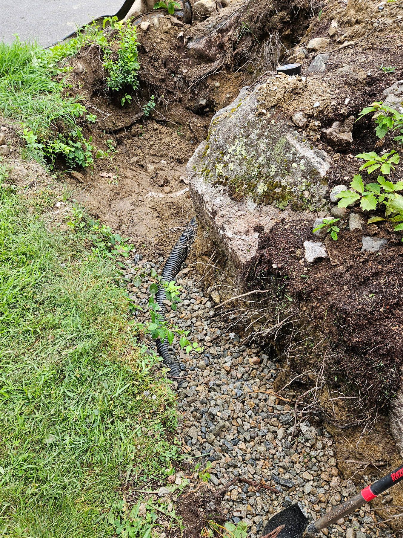 A trench with drainage pipe near a large rock, partly filled with gravel, adjacent to grass.