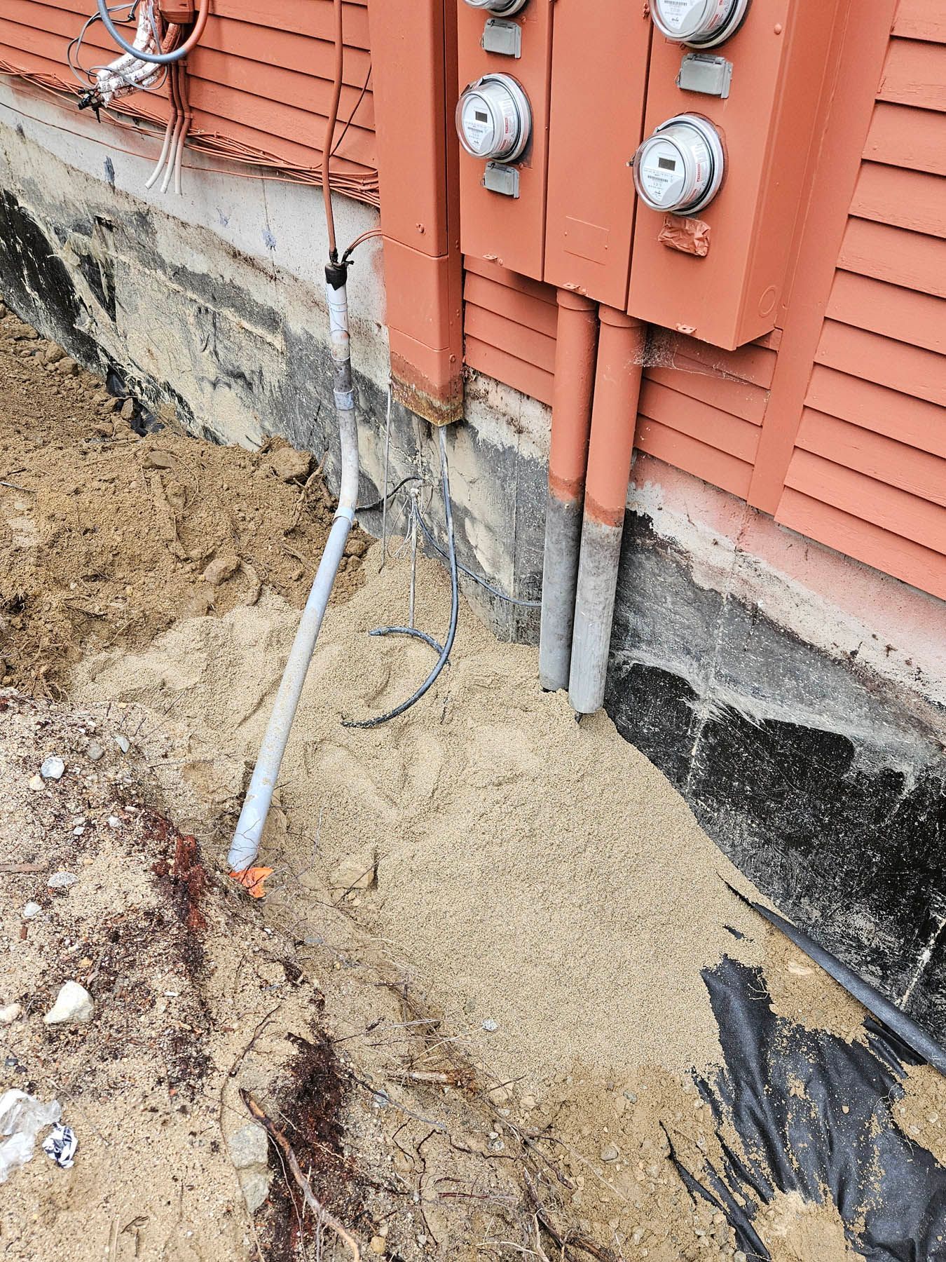 Exterior electrical meter boxes on a building with exposed foundations, a trench filled with wood chips and wiring.