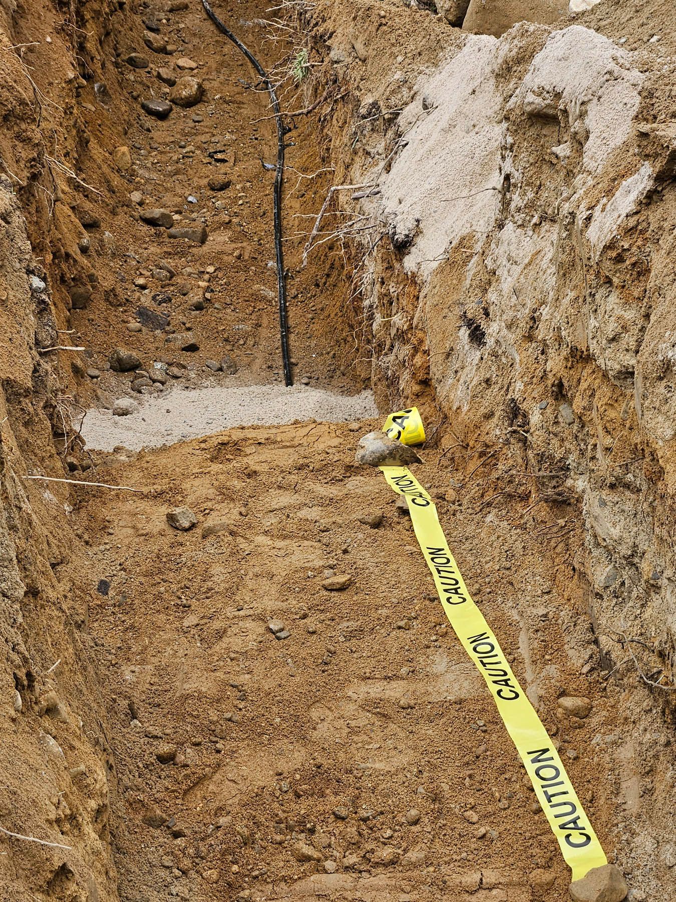 A narrow trench dug in earth; yellow caution tape laid along the bottom; a black pipe is visible.