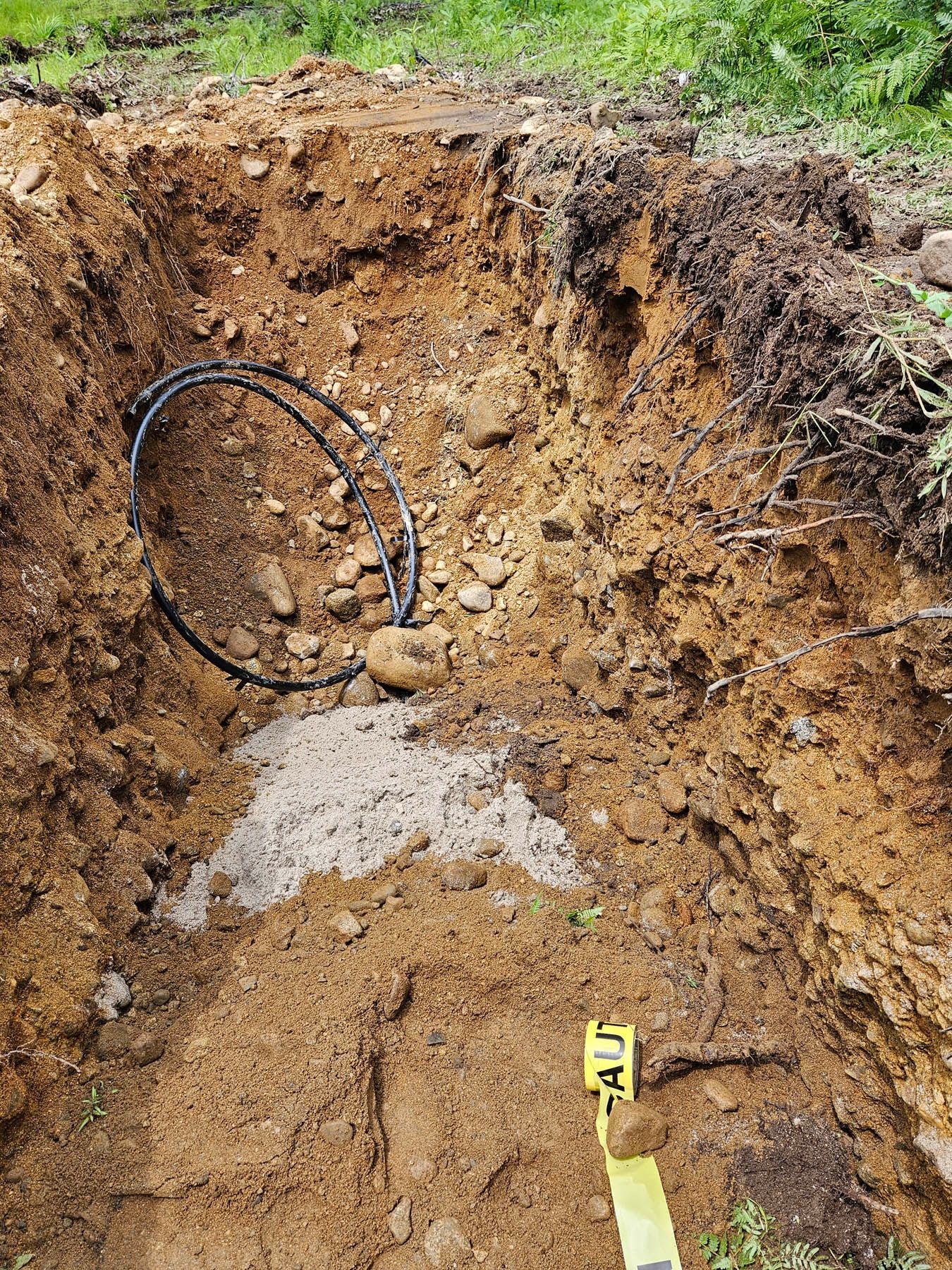 Excavated hole in soil, black coiled cable, gray cement, caution tape.