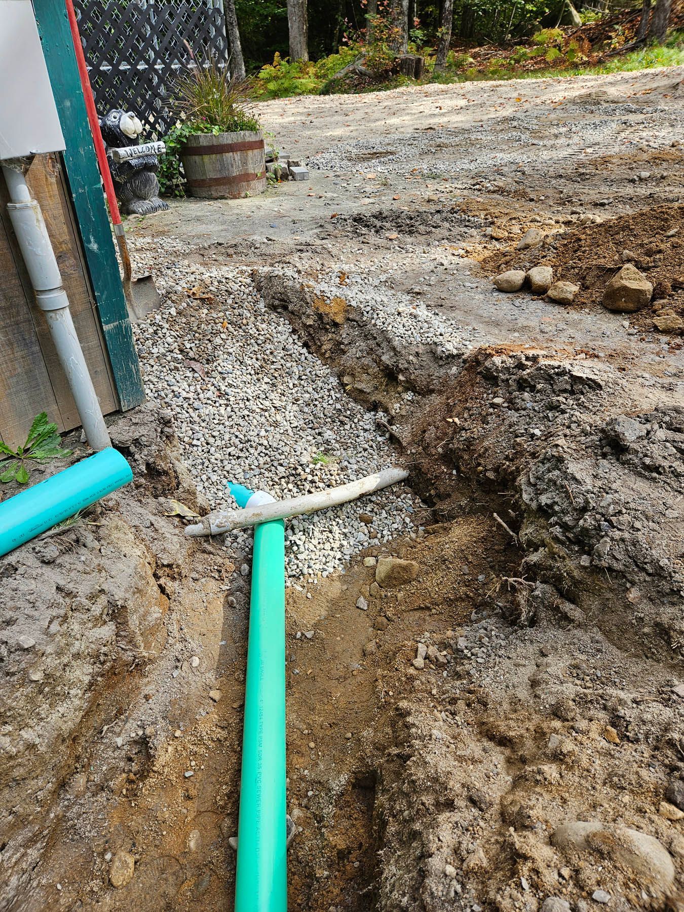 Trench dug for green pipe installation with gravel base, near building and dirt driveway.