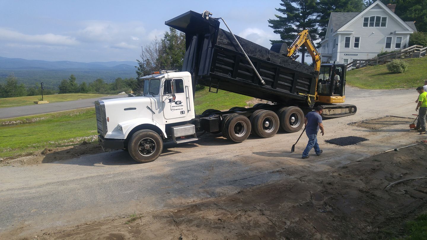 Dump truck being loaded with asphalt by an excavator. Workers and a house are in the background.