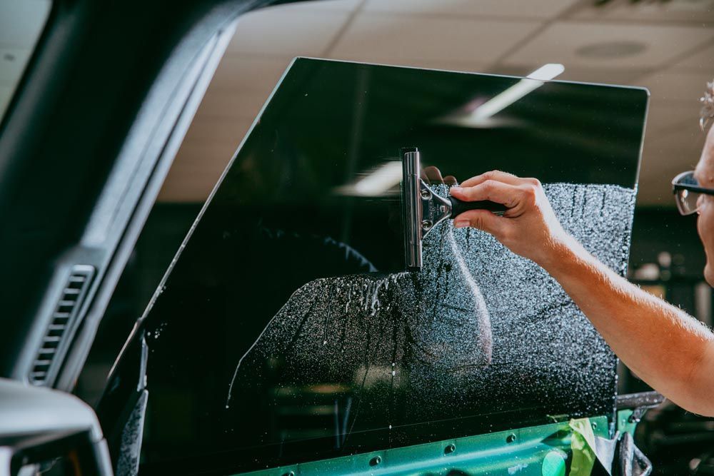 Person applying tint film to a car window with a squeegee. Inside a vehicle with green trim.