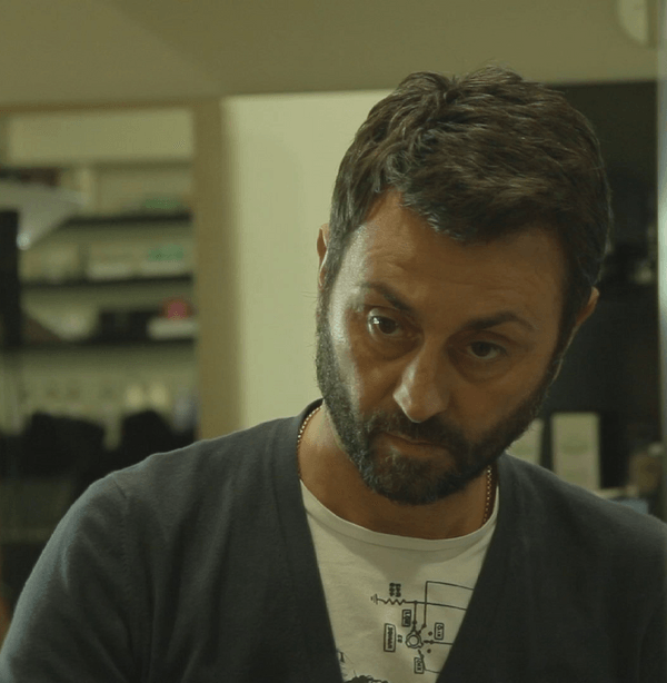 Uomo con barba e capelli scuri, che indossa un maglione blu sopra una maglietta bianca.