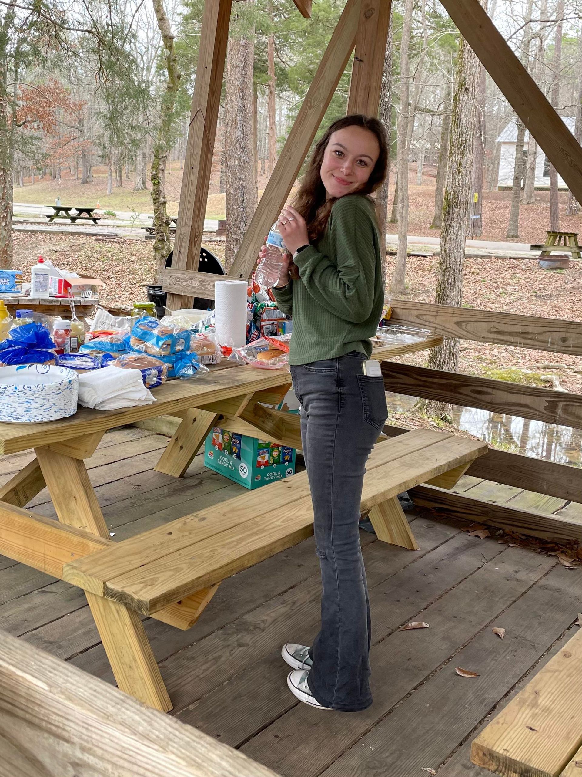 A woman is standing in front of a picnic table in the woods.