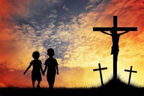 Two children are holding hands in front of a cross at sunset.