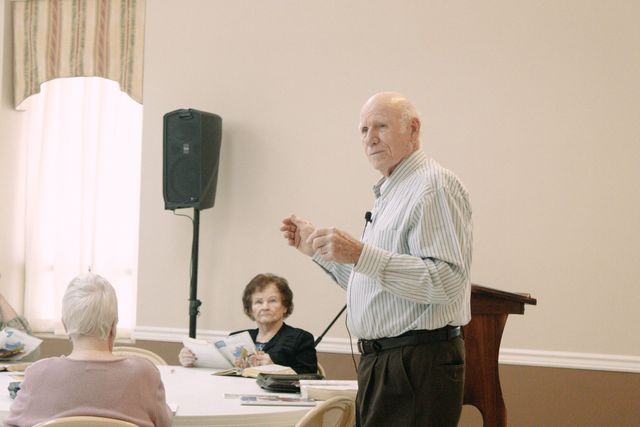 Man speaking at a podium in a meeting room while attendees sit and listen nearby