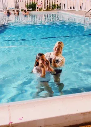 Two children standing in a blue swimming pool, one holding a phone while the other smiles nearby.