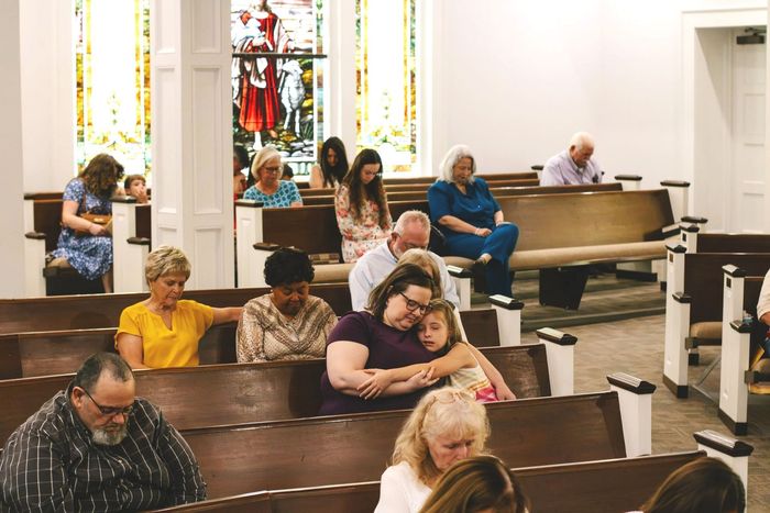 People seated in church pews during a quiet service, with stained glass windows and a pianist at the piano.