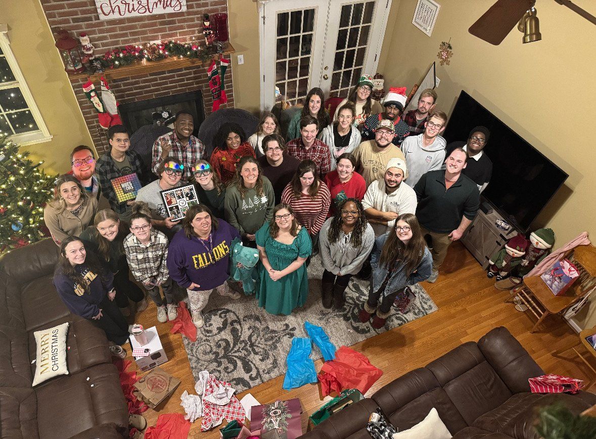 Large group posing in a living room, many holding wrapped gifts near a decorated fireplace and Christmas tree.