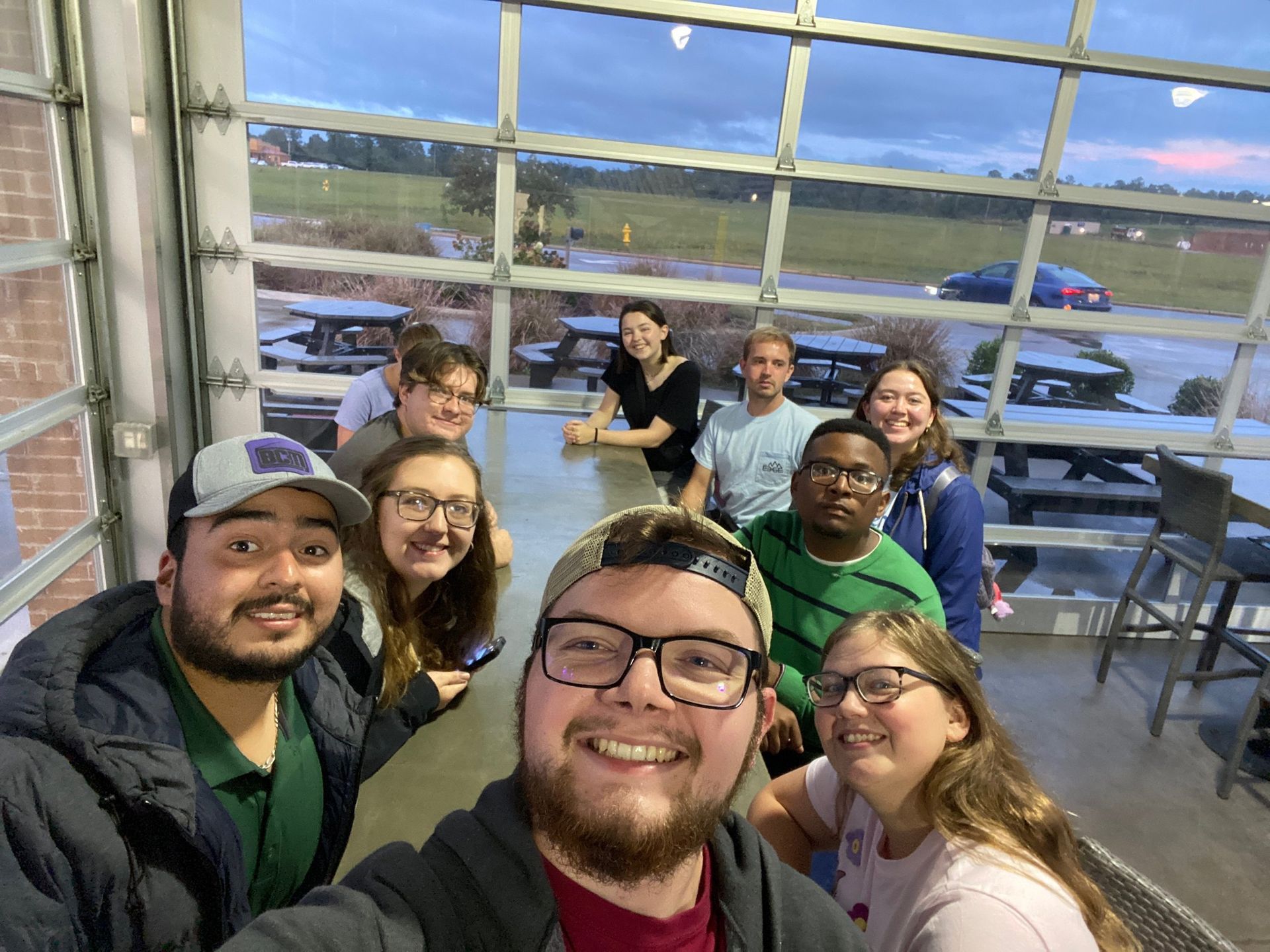 Smiling group selfie in a garage with open door, showing parked cars and green hills outside.