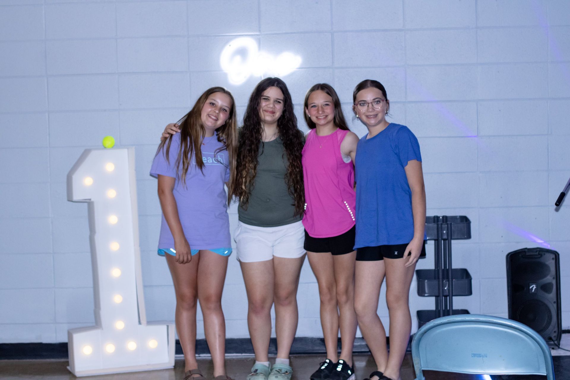 Four people posing indoors beside a large illuminated number 1 and speakers at a celebration.