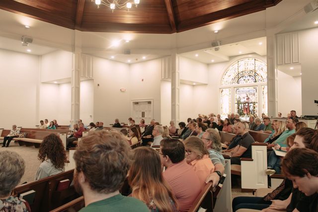 People seated in a church or auditorium during a gathering, with stained-glass windows and warm lighting.