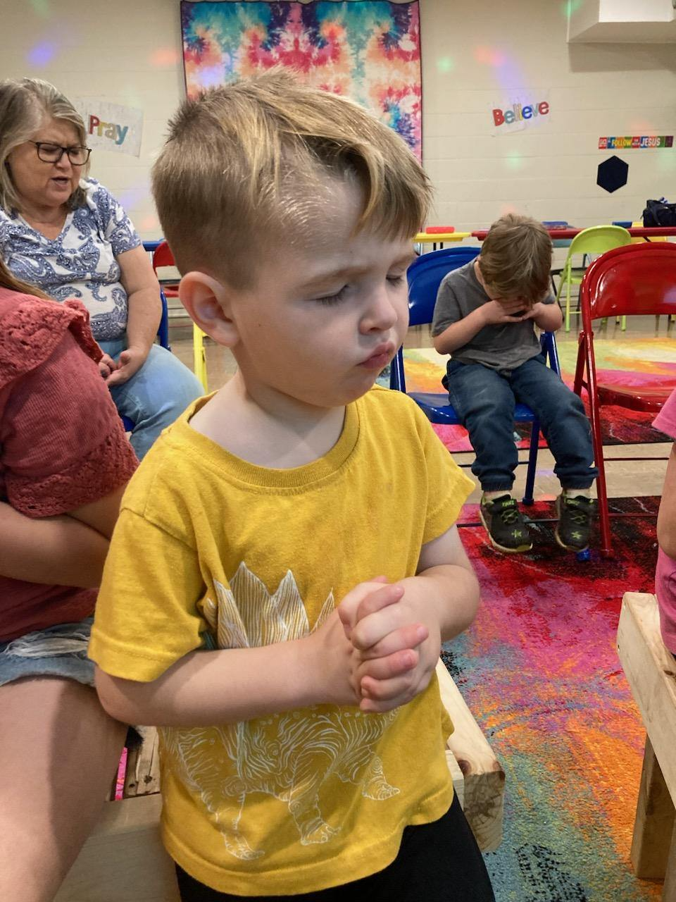 A young boy in a yellow shirt is praying in a classroom.