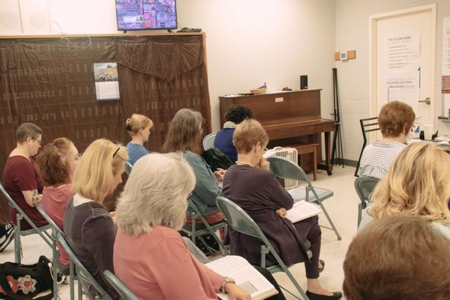 Audience seated in a room facing a piano and microphone stand, with a speaker at the front.