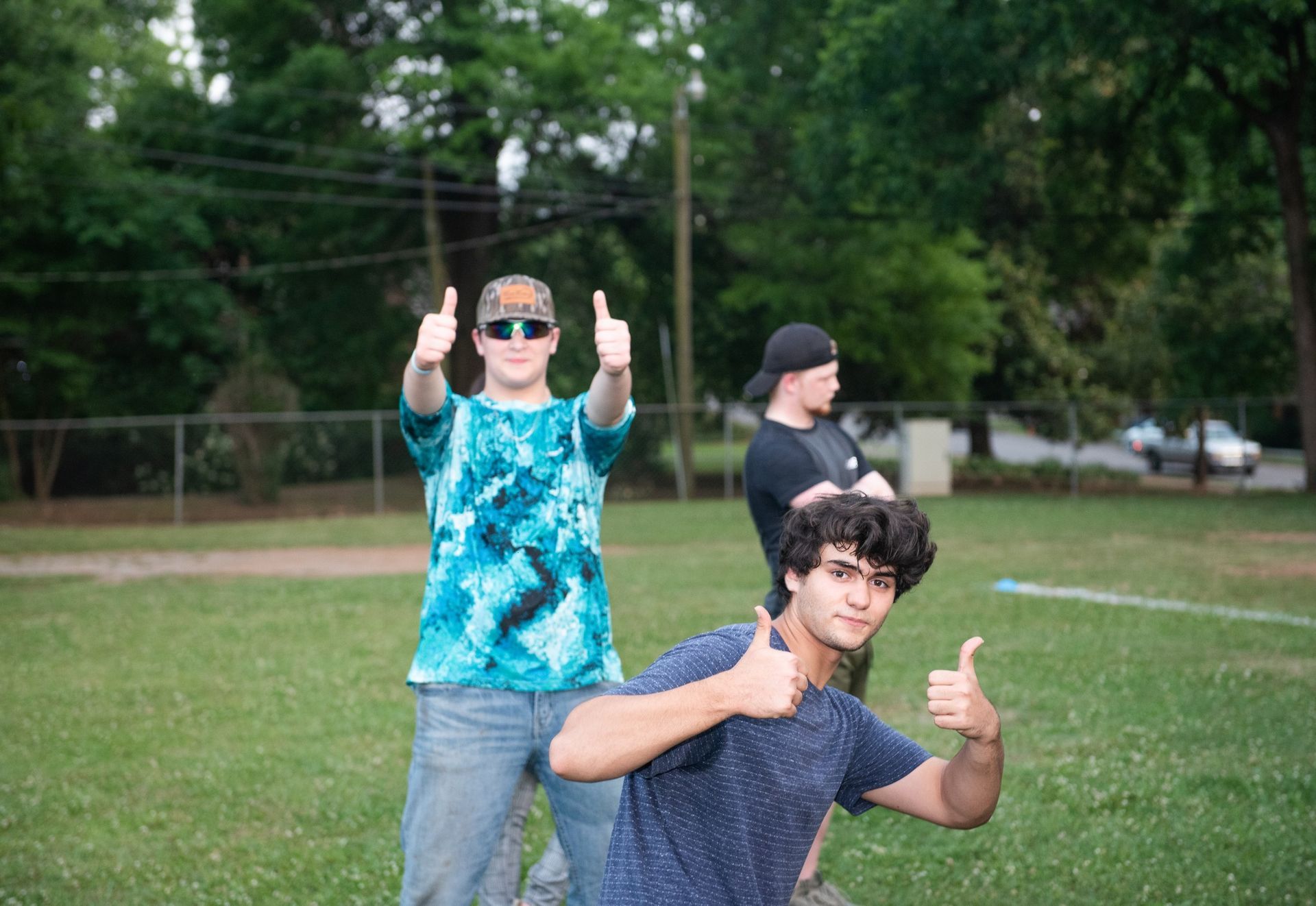 Three young men are giving a thumbs up in a field.