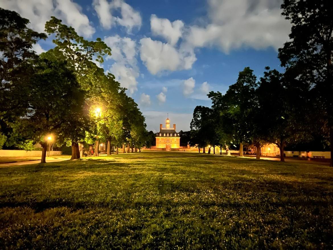 A long, grassy path lined with trees leads to a lit building at dusk under a cloudy sky.