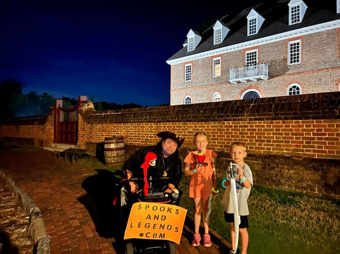 A costumed person in a scooter with two children on a brick path at night near a historic building.