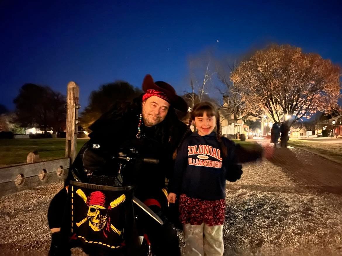 A person in pirate costume with a child, posing outdoors at night. The child smiles and waves.