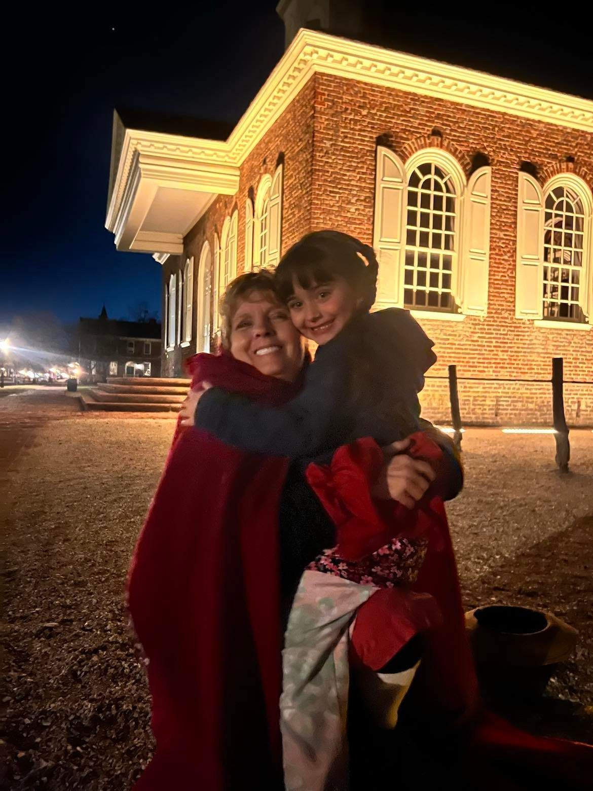Woman hugging a child in front of a brick building at night. They wear red and dark clothing, smiling.
