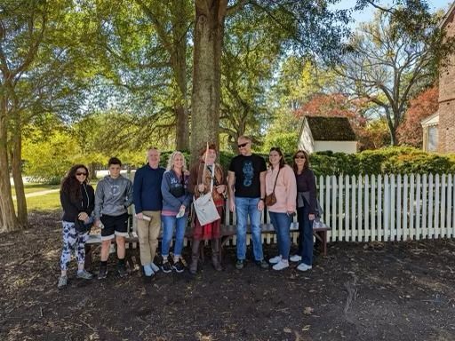 Group of people standing near a tree and a white picket fence in a park setting.