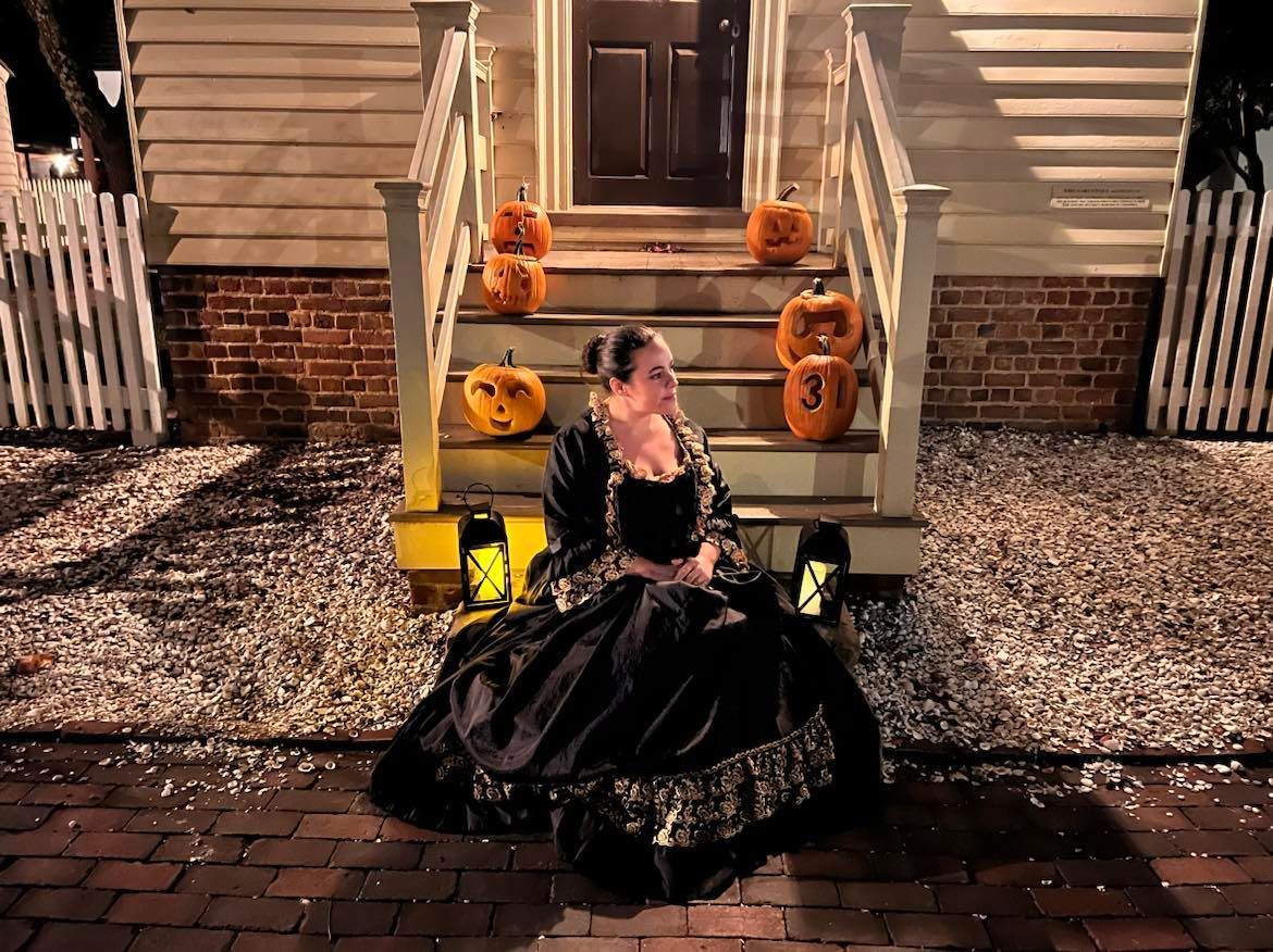 Woman in black gown sits on steps with carved pumpkins, lanterns, and a white fence.