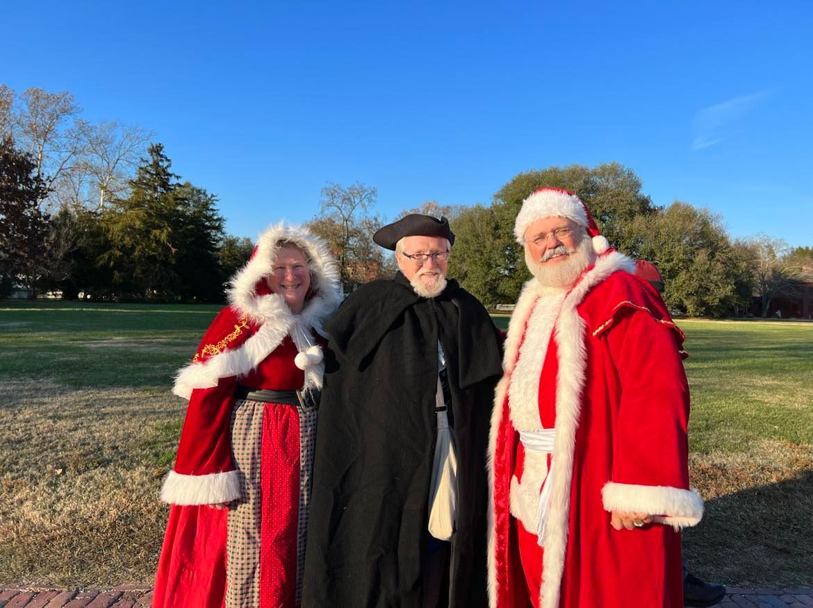 Three costumed figures outdoors: one in black, two in red Santa suits, smiling.