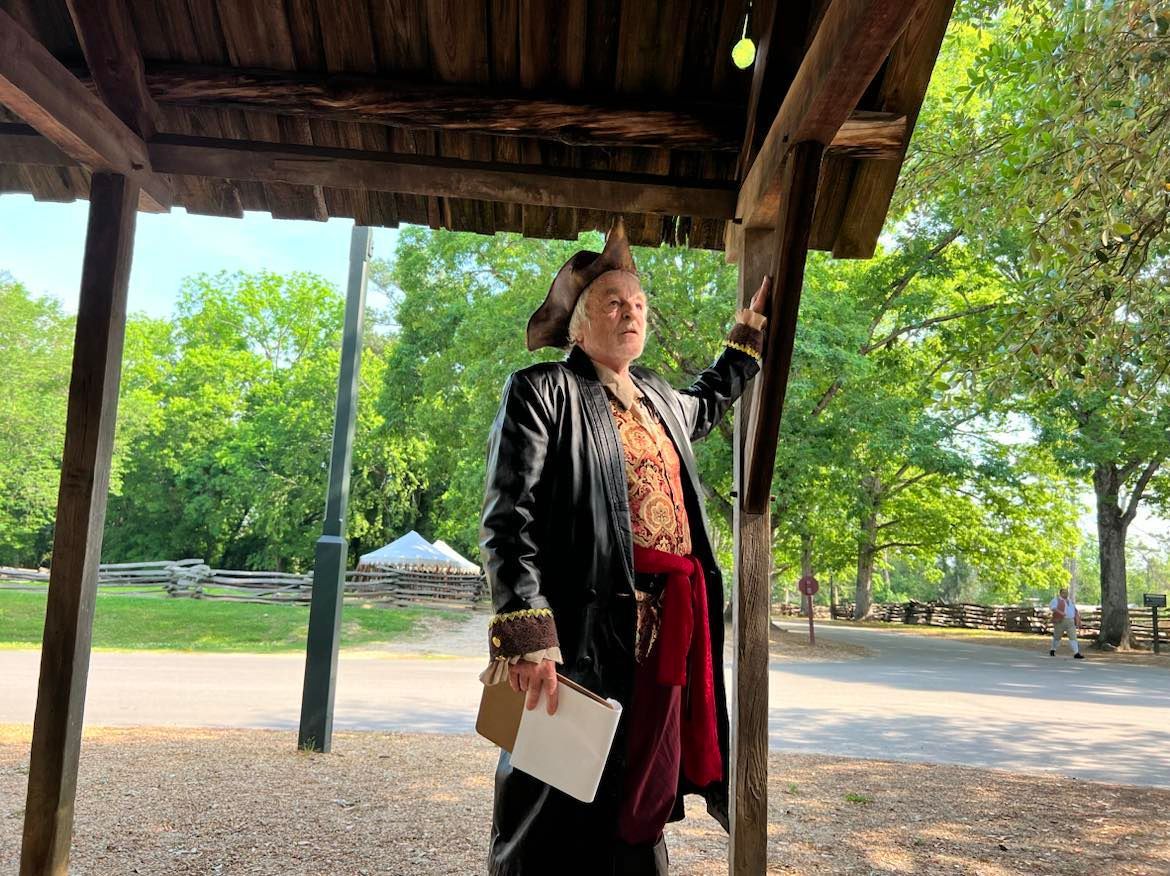 Person in period clothing pointing upward under a wooden structure; park setting with trees.