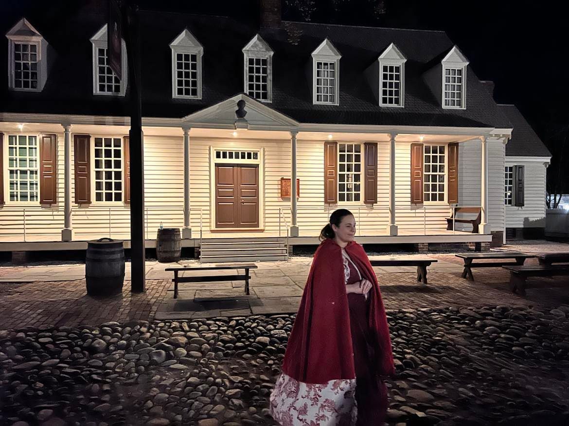 Woman in red cape stands before a historic building at night, cobblestone ground.