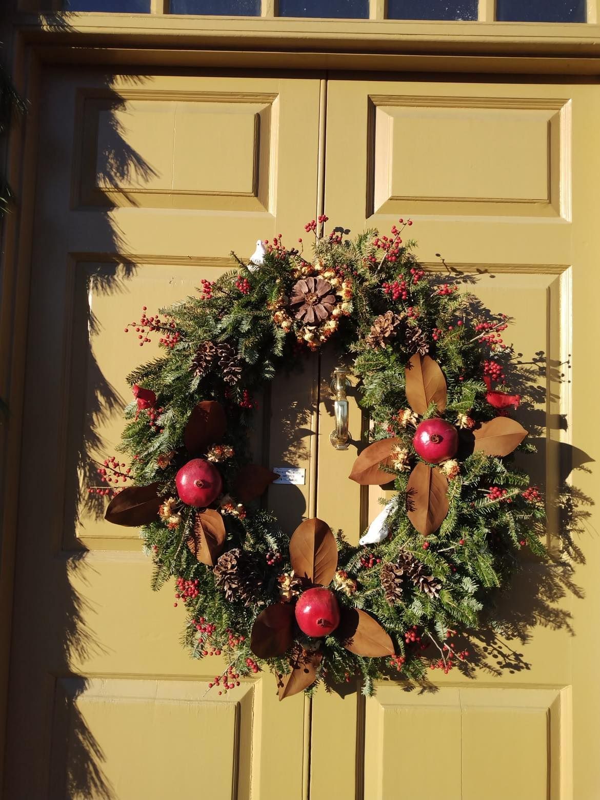 Wreath on yellow door: Green branches, red berries, brown leaves, pinecones, and red round ornaments.