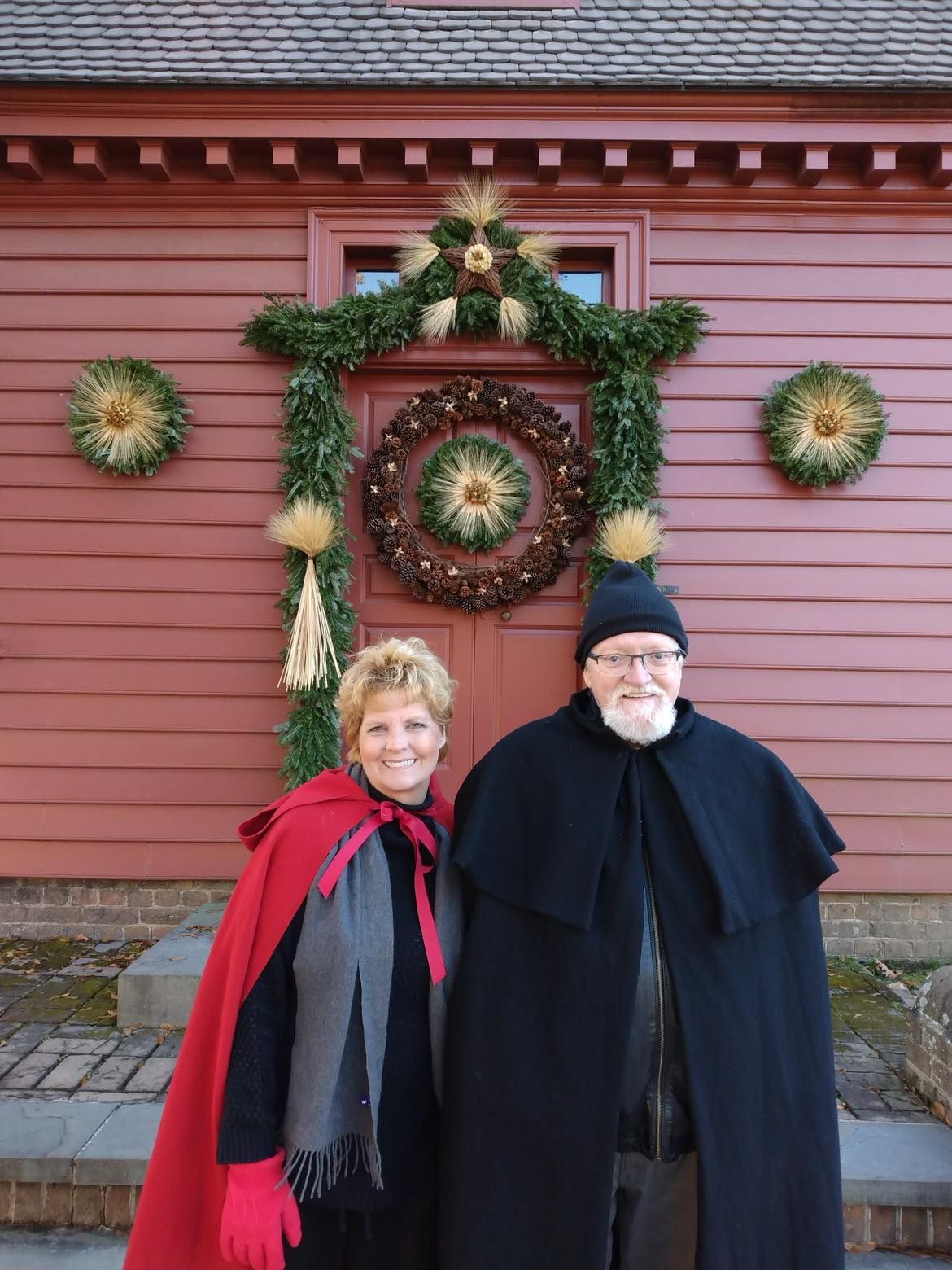 Couple in capes poses in front of a red building with holiday decorations.