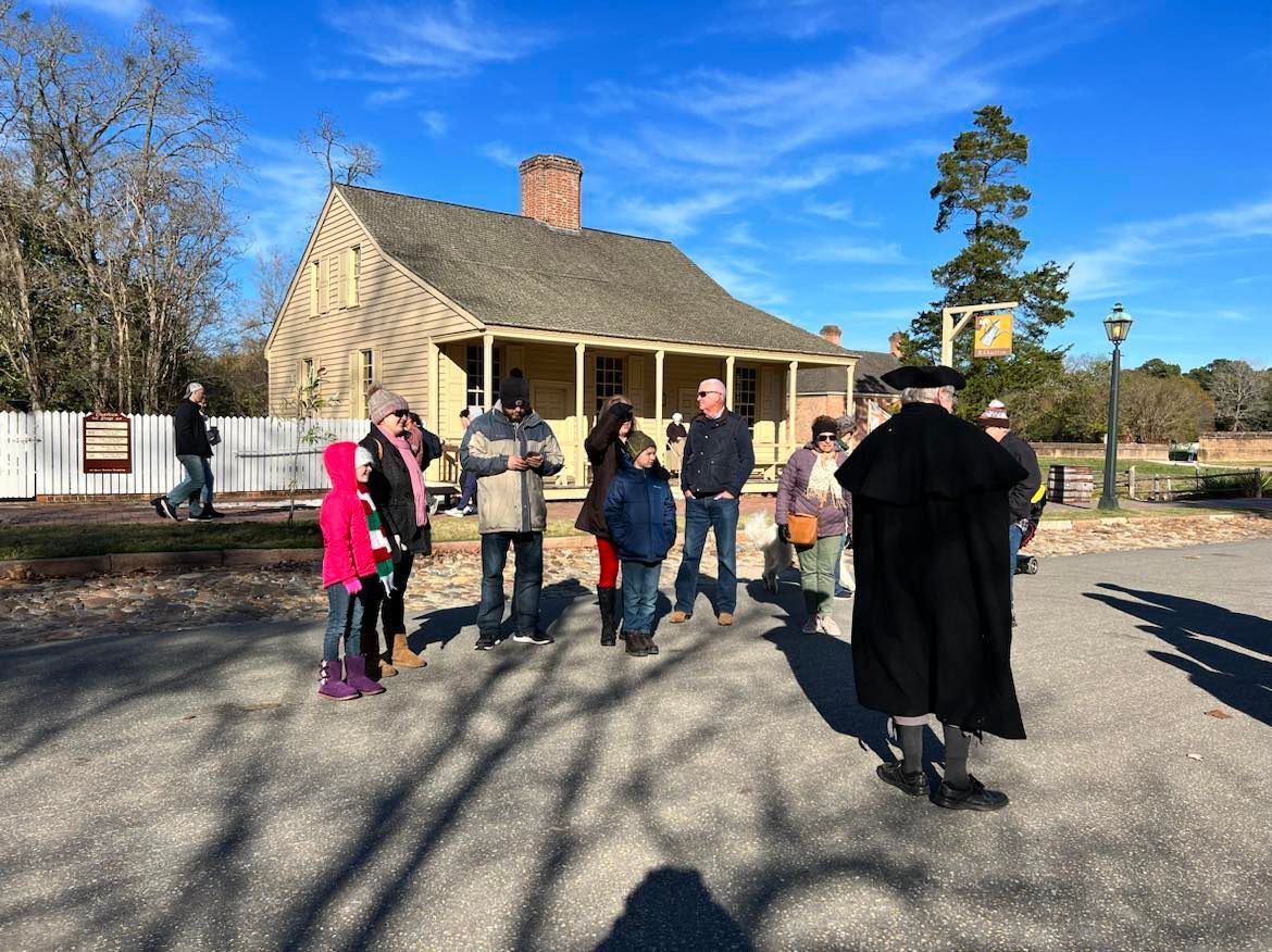 A group of people listens to a man in colonial attire outside a yellow building. Blue sky.