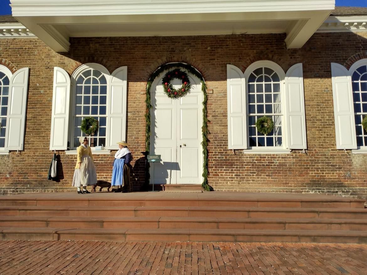 Brick building facade with white arched windows and door. Two figures in period clothing stand by door with wreath.