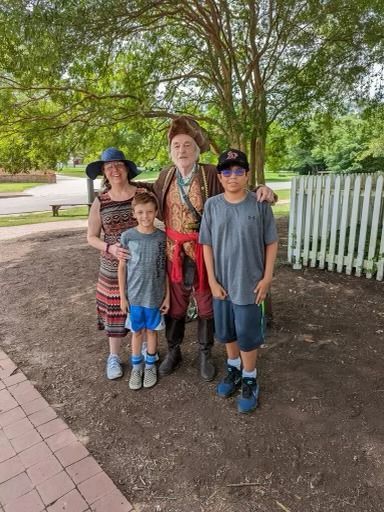 Family poses with a costumed historical figure near a white fence, under a tree.