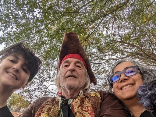 Three people smiling for a selfie outdoors; one wears a pirate hat and costume, trees overhead.