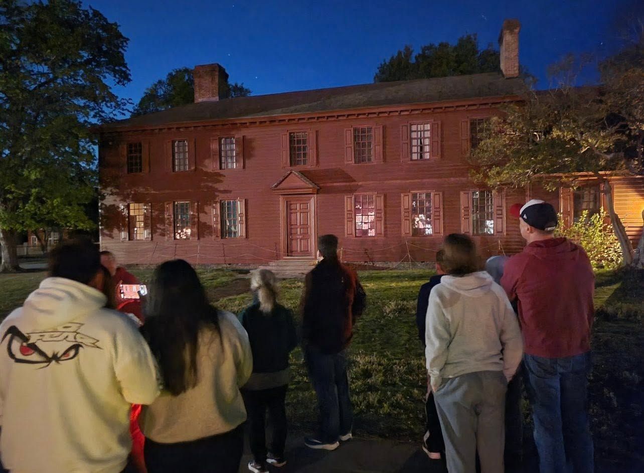 Group of people viewing a lit brick mansion at dusk from the lawn