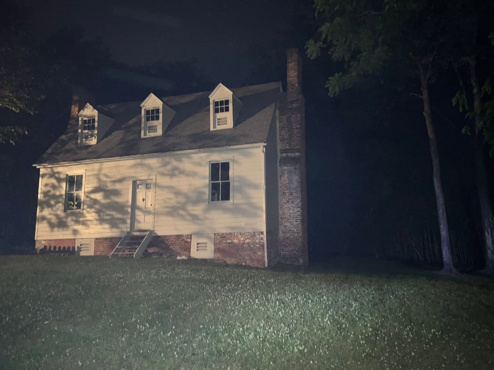 Night view of a white house with dormer windows, lit by a spotlight beside tall dark trees and a grassy lawn