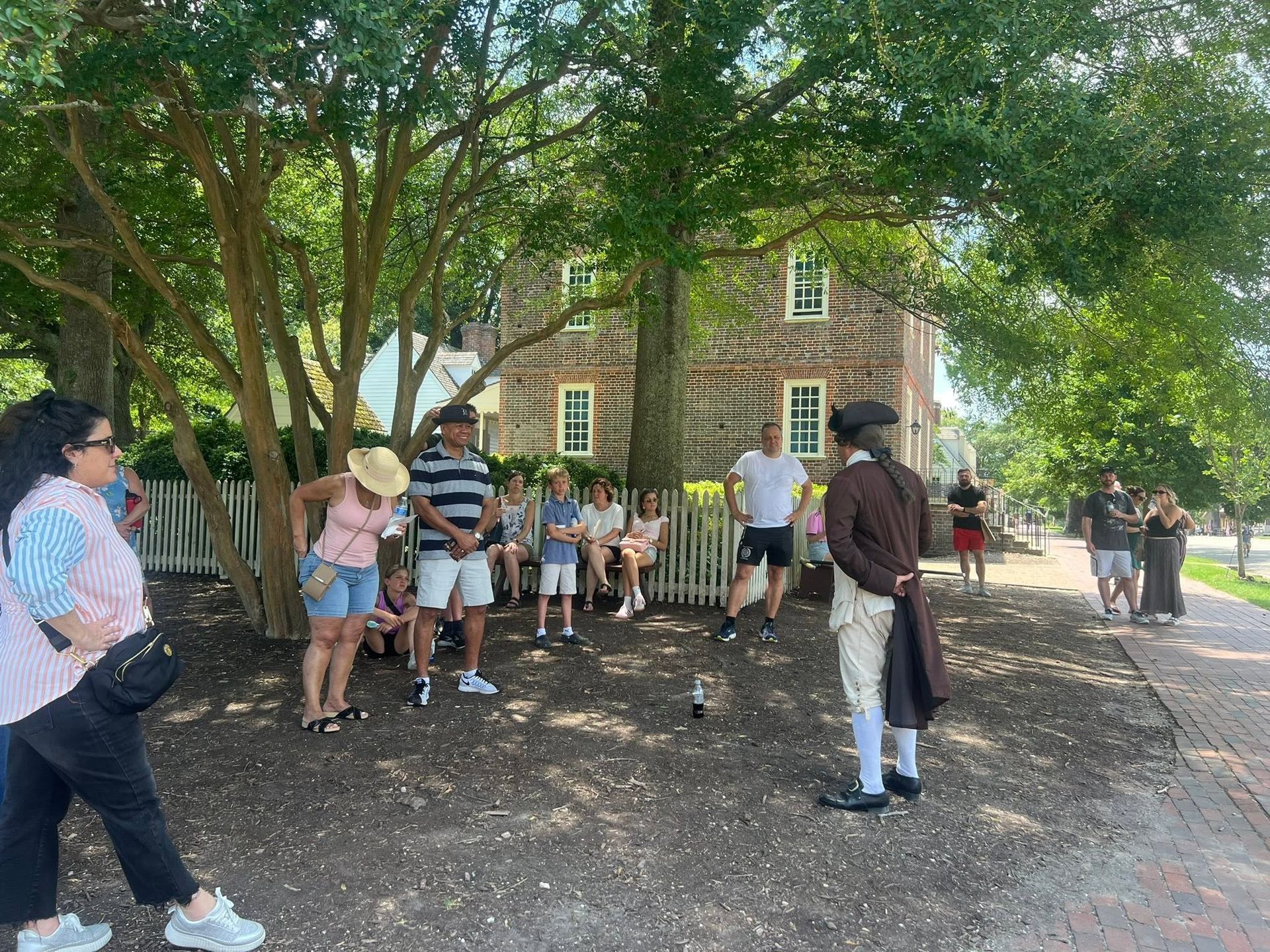 People gathered under large trees outside a brick building, some standing and chatting, others seated on benches.