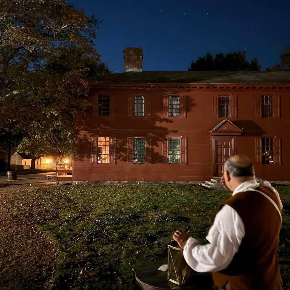Man in vest looking at a lit red-brick house at night under a dark blue sky