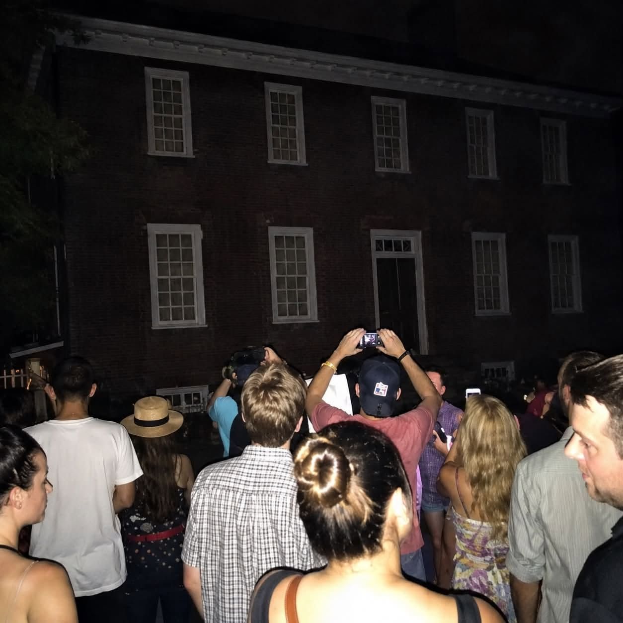 Crowd gathered at night facing a dark brick building with lit windows, some people holding up phones.