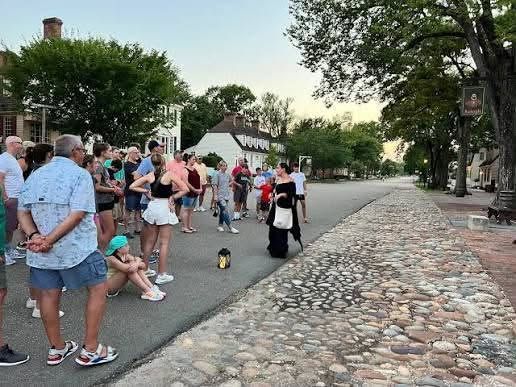 People gathered along a road beside a stone wall and trees, watching something off to the right.