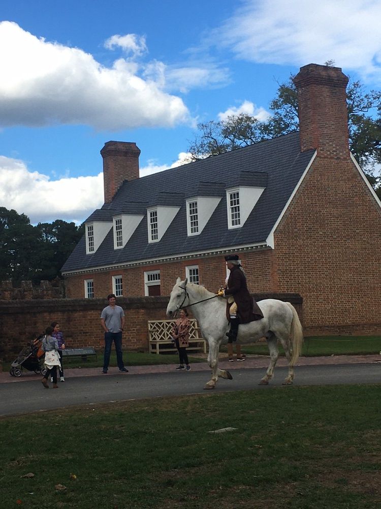 Person in historical attire riding a white horse near a brick building. People watch.