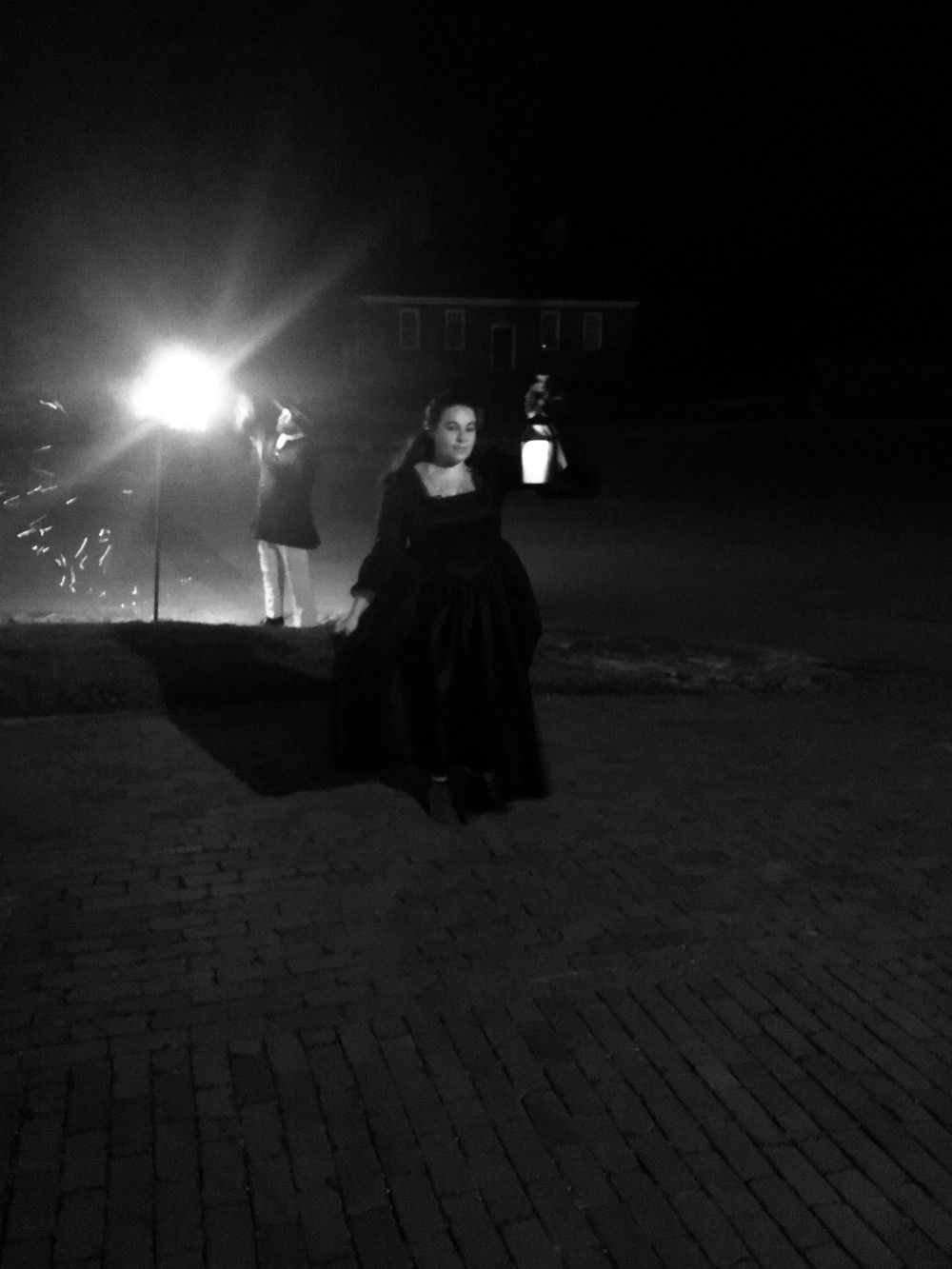 Woman in dark dress holds lantern at night, a second figure stands near bright light.