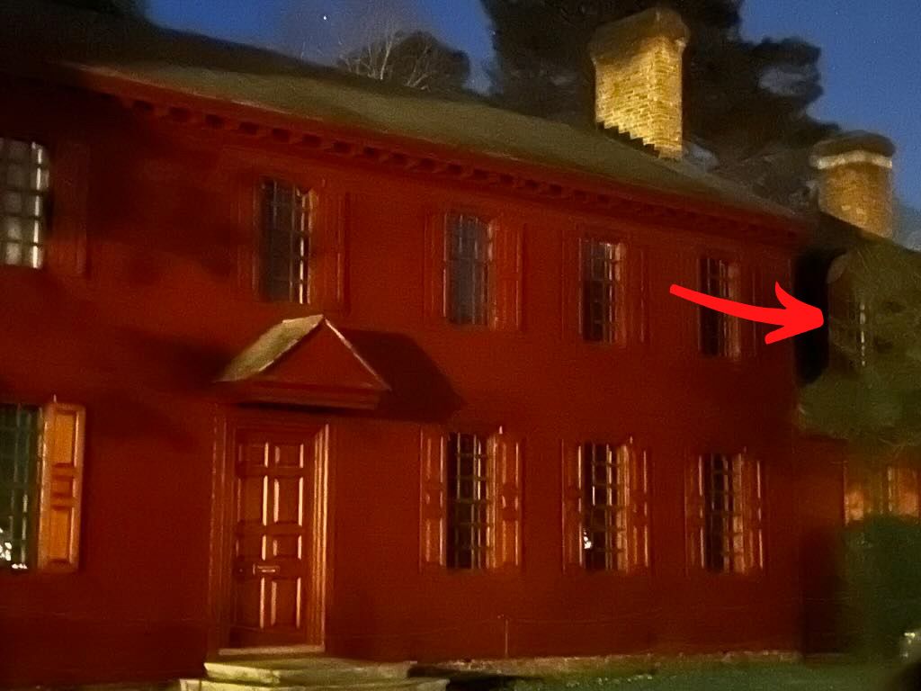 Red, two-story house with shutters, brick chimneys, and an arrow pointing to a dark corner, possibly a side entrance.