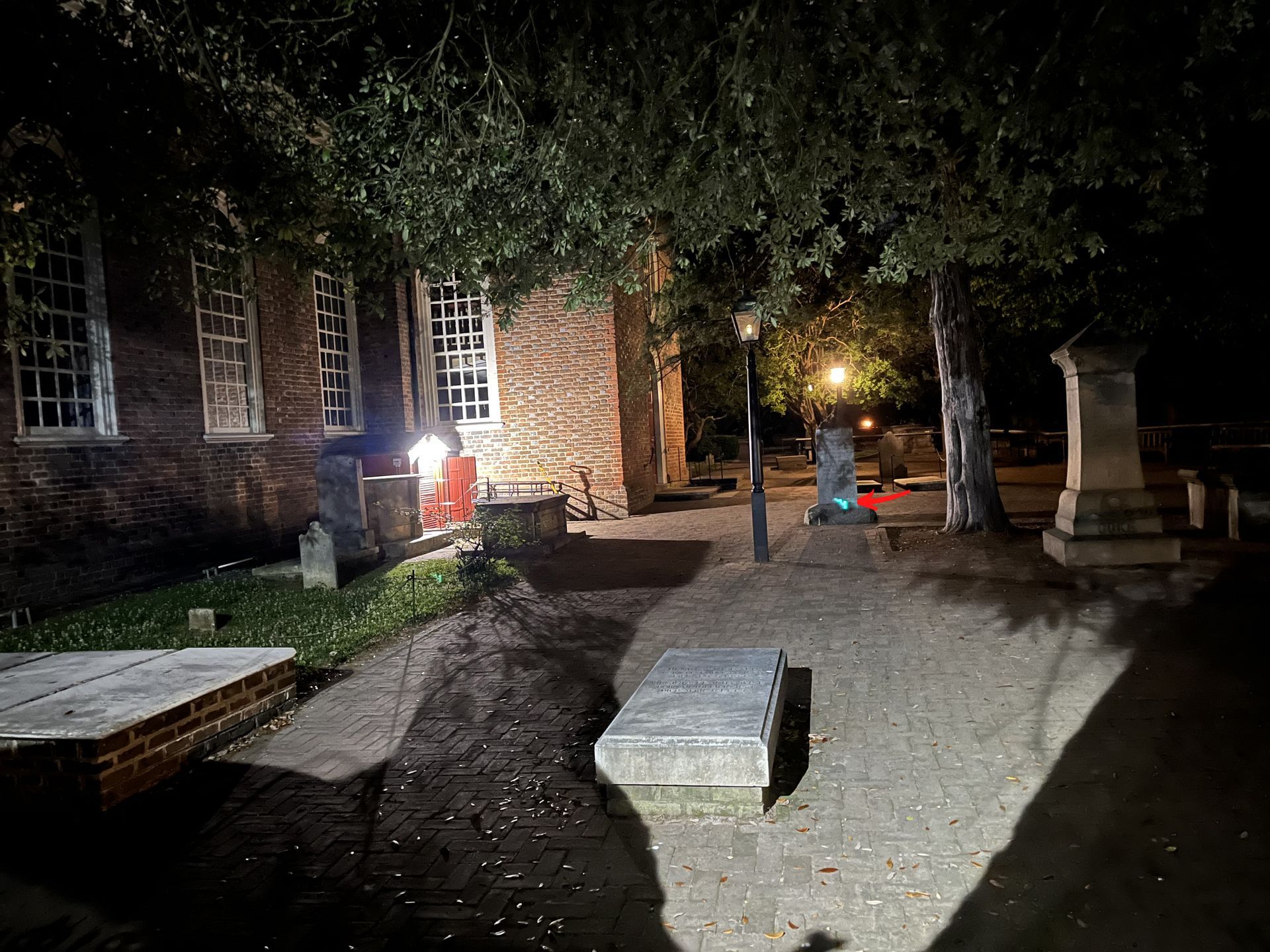Night shot of a brick church and graveyard with tombstones under tree branches and a street lamp.