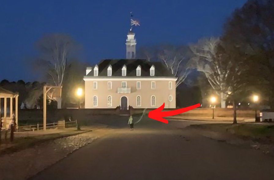 Two people walk down a road at night toward a lit building with a flag; streetlights and trees line the road.