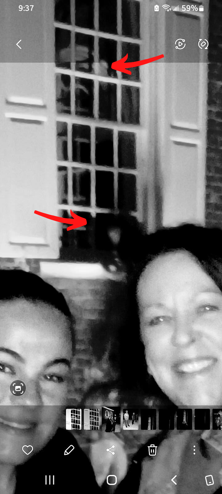 Two women smiling in front of a window. A blurred figure is visible behind the window's panes.
